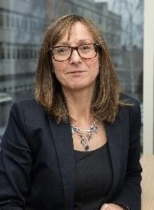 Ann Carruthers, a white woman with brown hair and glasses, wearing a black suit, looking at the camera and smiling