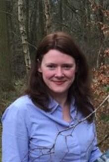 Donna Tavernor, a white woman with long dark brown hair, wearing a blue shirt, standing in woodland, looking at the camera and smiling