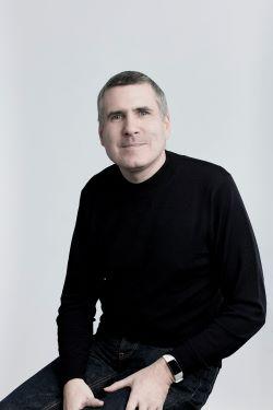 Giles Perkins, a white man with very short grey hair, wearing a black jumper and trousers, sitting on a stool and looking at the camera