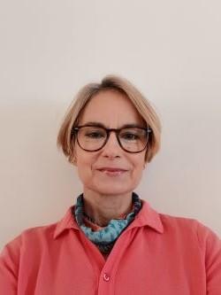 Sonia Hansen, a white woman, with short blonde hair and large glasses, wearing a salmon pink top and looking at the camera