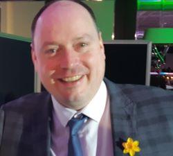 Rob Shoebridge, a white man, bald, wearing a dark suit, white shirt and blue tie, smiling at the camera