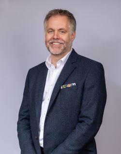 Shaun Helman, a white man with short grey hair and a full beard, wearing a dark suit and white shirt, smiling at the camera