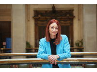 Amy Harhoff, a white woman with red hair, black top and bright blue jacket leaning on a rail