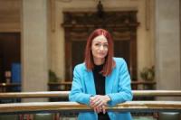 Amy Harhoff, a white woman with red hair, black top and bright blue jacket leaning on a rail