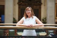 Ann Carruthers, a woman with brown hair and glasses, wearing a sleeveless white lace top, leaning on a balcony, looking at the camera and smiling