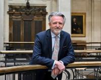 Anthony Payne, a white man in his late fifties, with short grey hair and a beard, wearing a dark blue suit, pale blue shirt and light-coloured tie, leaning on a balcony, looking at the camera