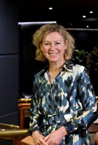 Hannah Bartram, a white woman in her early fifties, with short blond curly hair, wearing a grey and white patterned dress, looking at the camera and smiling.