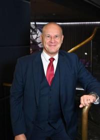 Mark Stevens, a white man in his fifties, bald, wearing a dark three-piece suit, white shirt and red tie, looking at the camera and smiling.