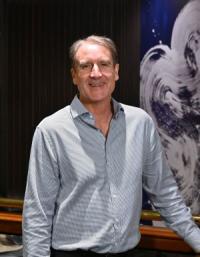 Neil Gibson, a white man in his late fifties, with short grey hair, wearing a pale blue shirt looking at the camera and smiling.