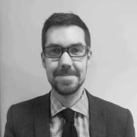 Mickey Green, a man in his 40s with short dark hair and a beard, wearing rectangular glasses, a checked shirt and tie. Photo is in black and white.
