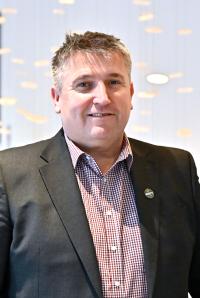 Mark Jackson, a white man in his forties with very short dark hair, wearing a dark suit jacket and red/white checked shirt.